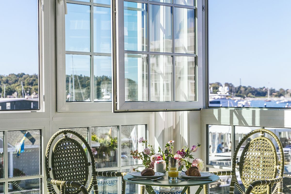 A sunlit room featuring a small table set with food, two chairs, and a large window with a scenic harbor view outside.
