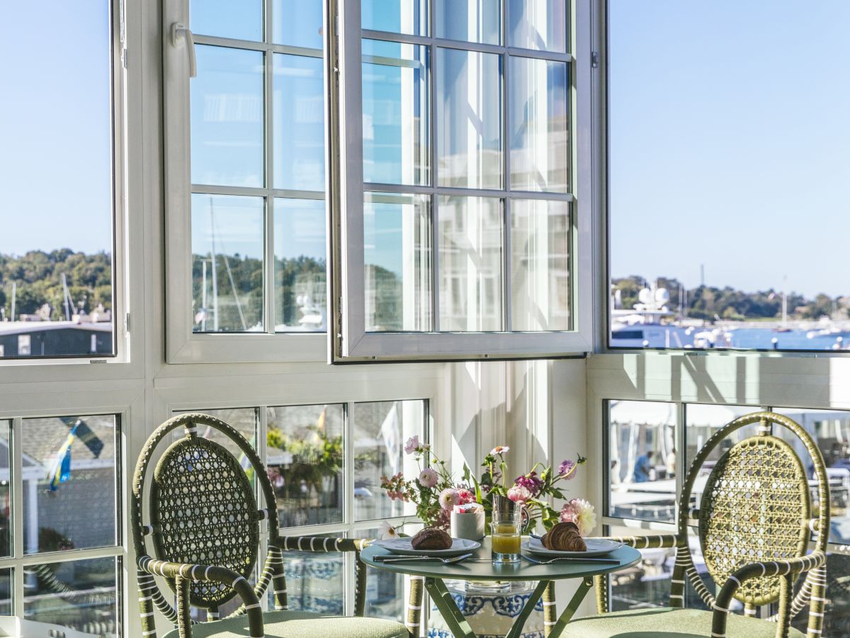 A sunlit room featuring a small table set with food, two chairs, and a large window with a scenic harbor view outside.