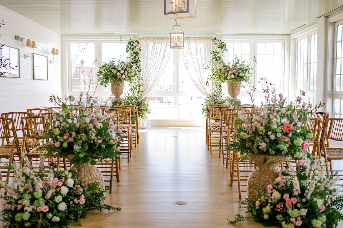 This image features an elegantly decorated indoor wedding venue with floral arrangements and wooden chairs lining the aisle.