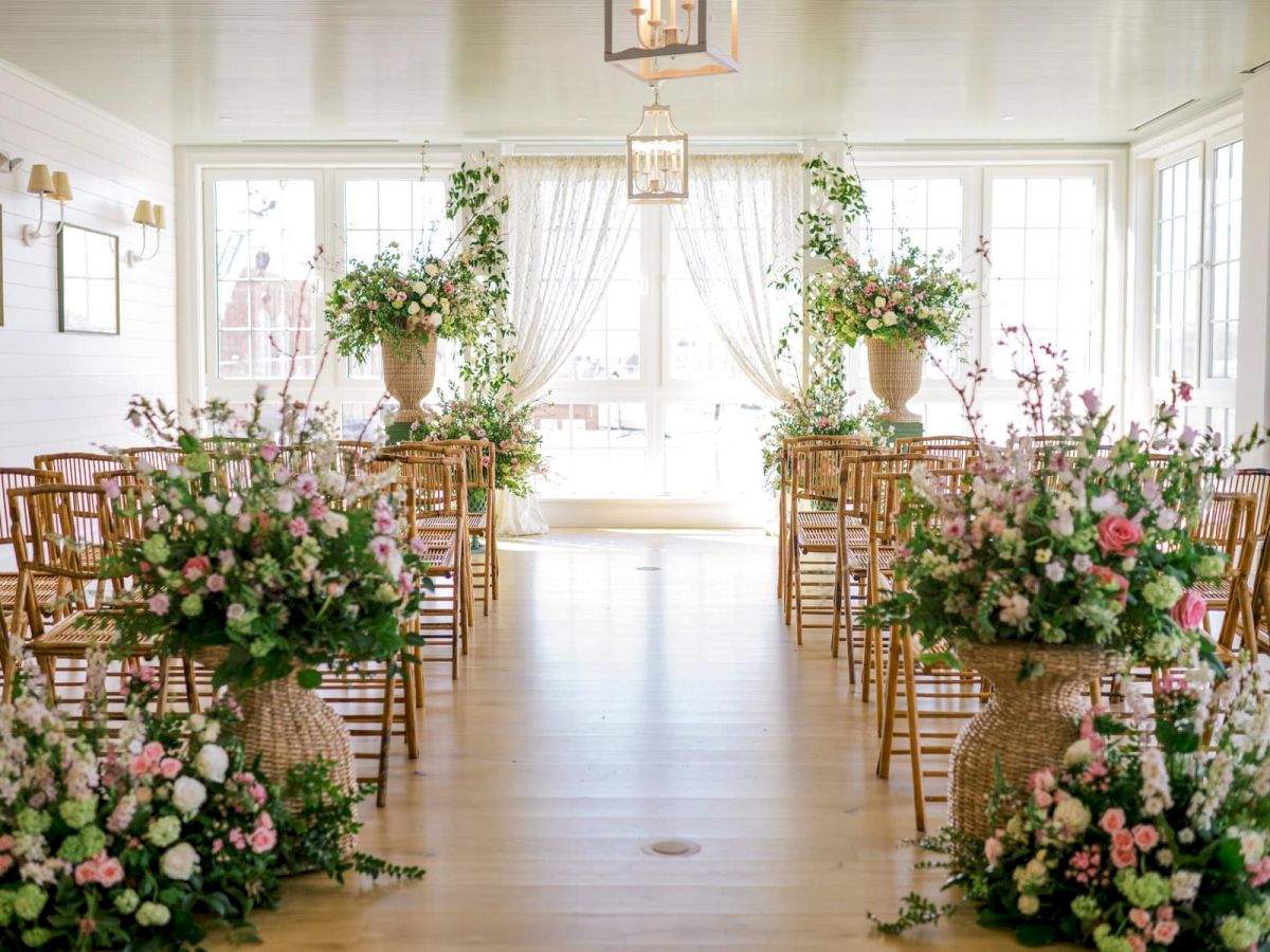 This image features an elegantly decorated indoor wedding venue with floral arrangements and wooden chairs lining the aisle.
