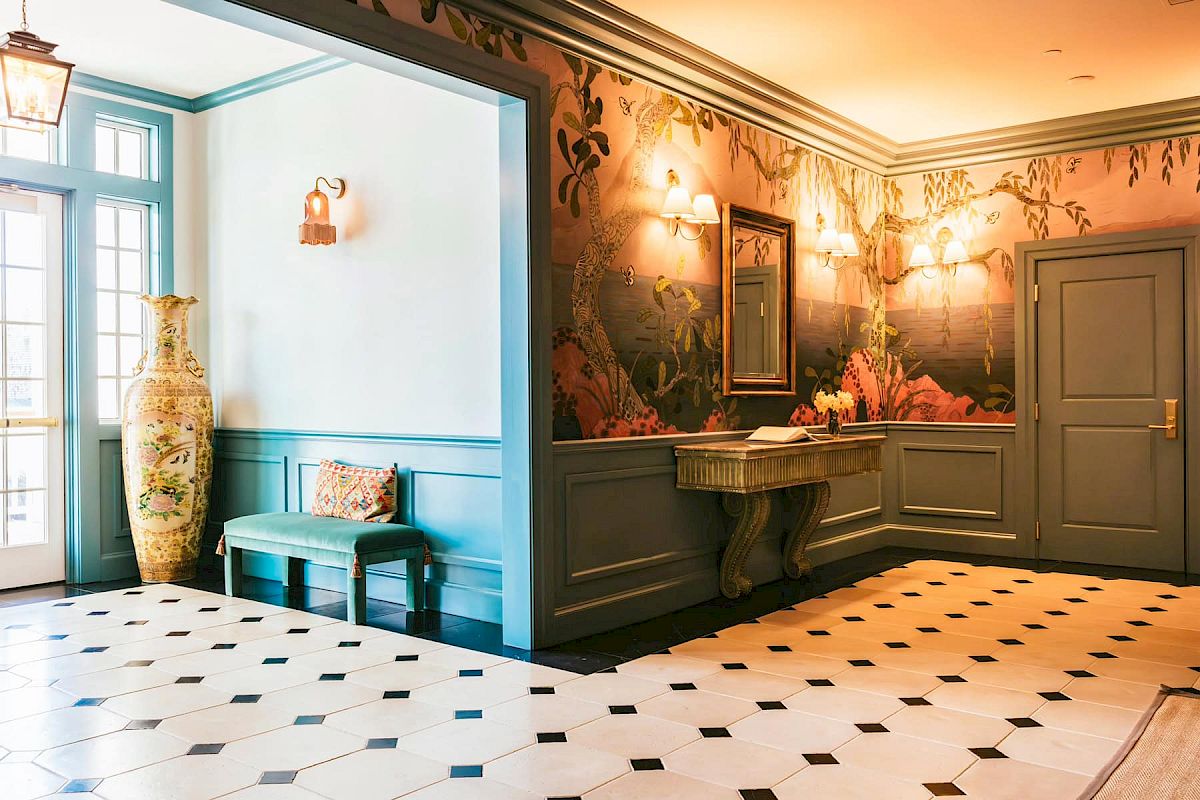 The front hall featuring elegant wallpaper, a green bench with cushions, a large vase, a mirror above a table, and soft lighting.