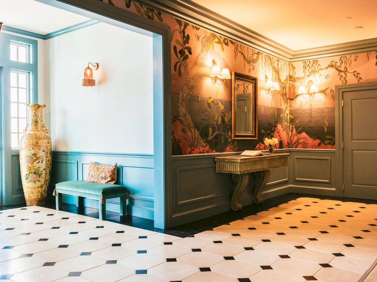 The front hall featuring elegant wallpaper, a green bench with cushions, a large vase, a mirror above a table, and soft lighting.