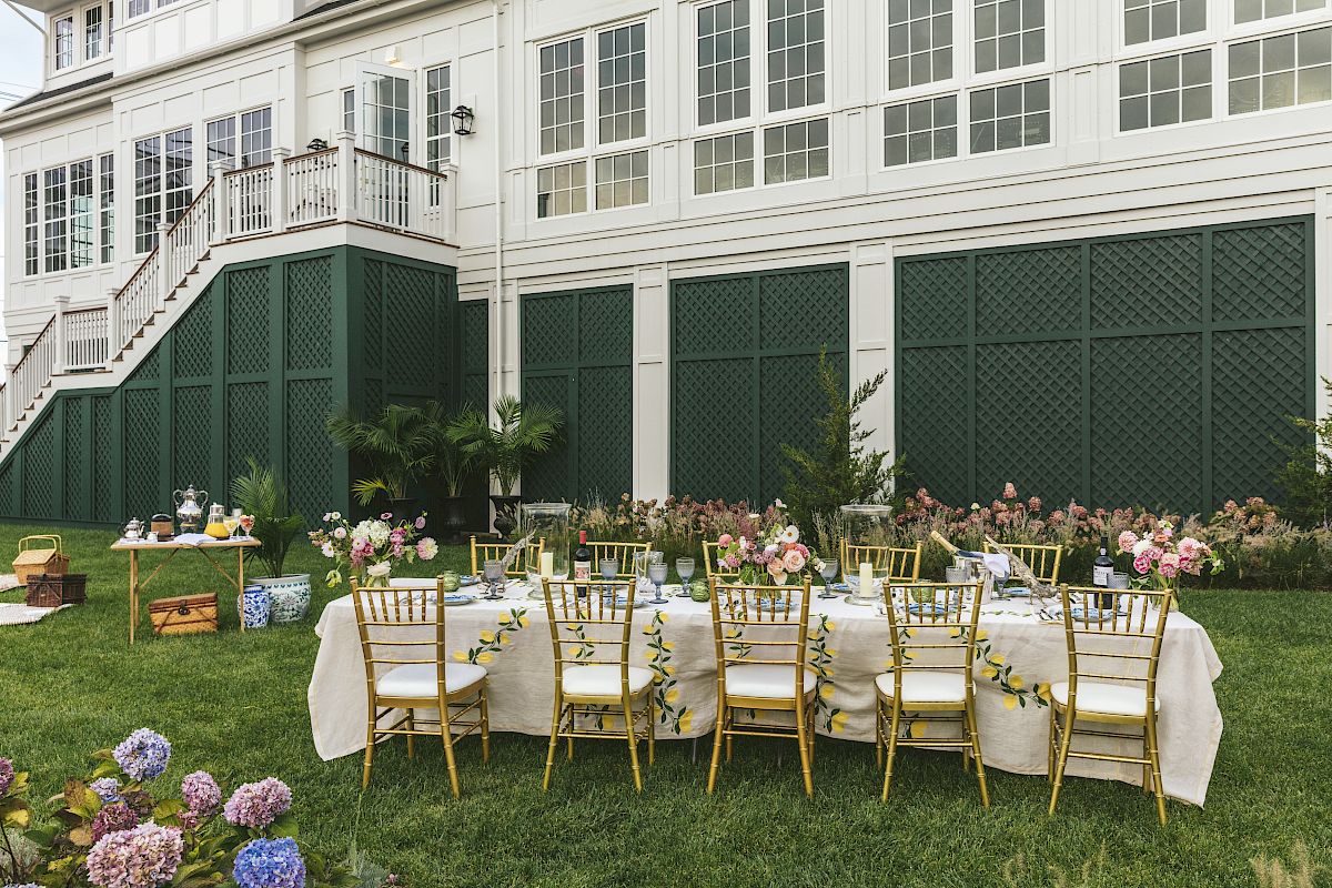 An outdoor setting features a long table with floral decorations and gold chairs, set against a white building with a staircase.