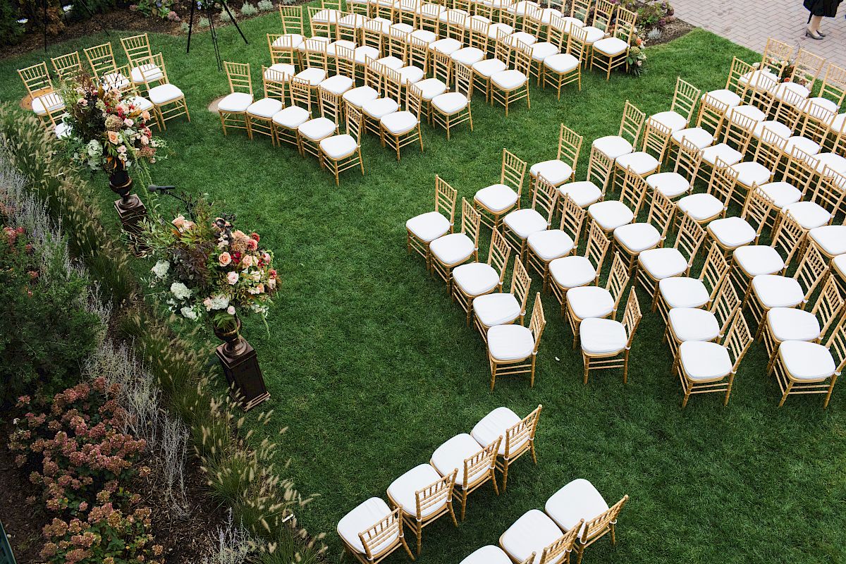 An outdoor event setup with rows of white-cushioned chairs arranged on the lawn surrounded by flowers and greenery.