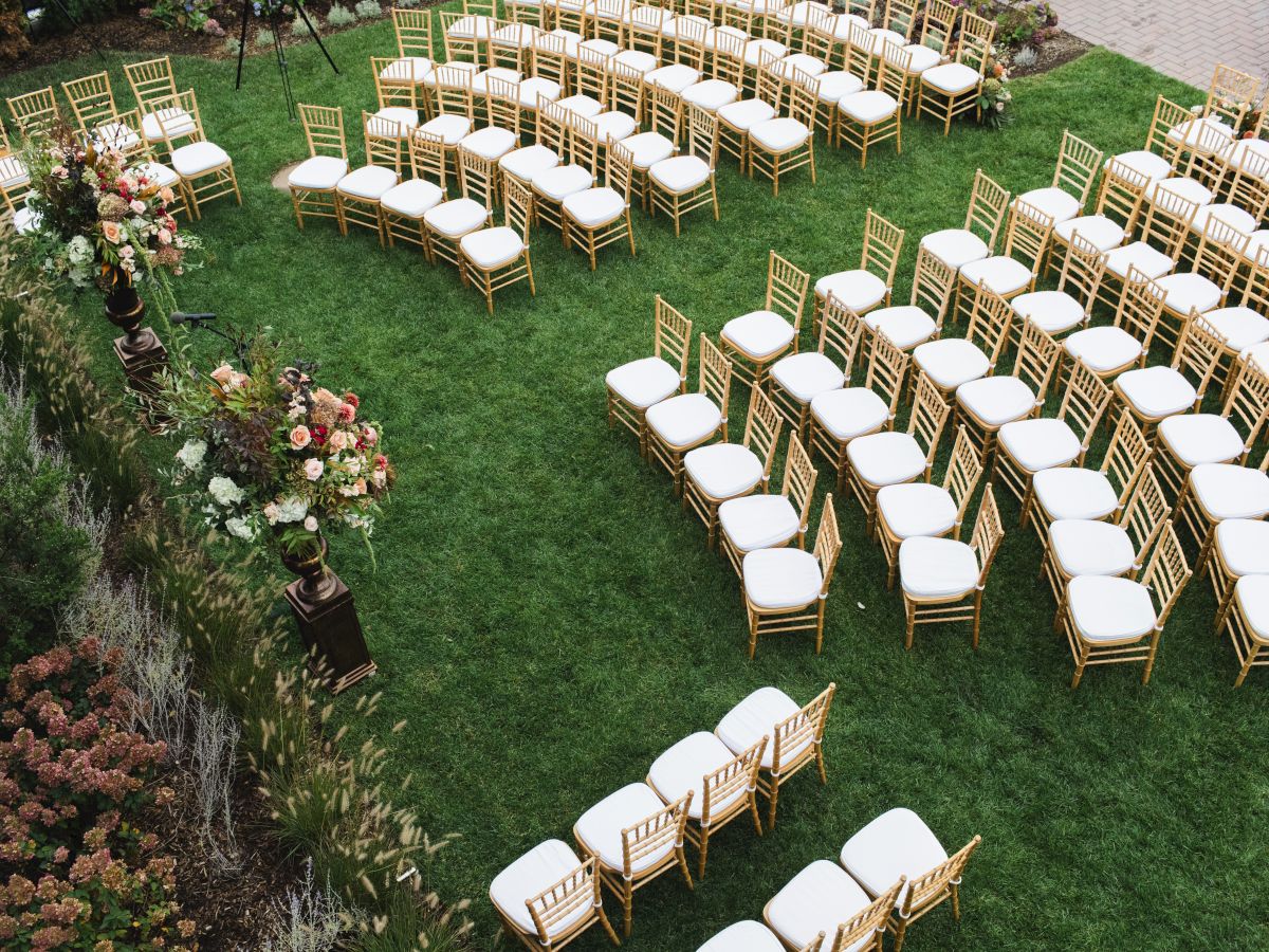 An outdoor event setup with rows of white-cushioned chairs arranged on the lawn surrounded by flowers and greenery.