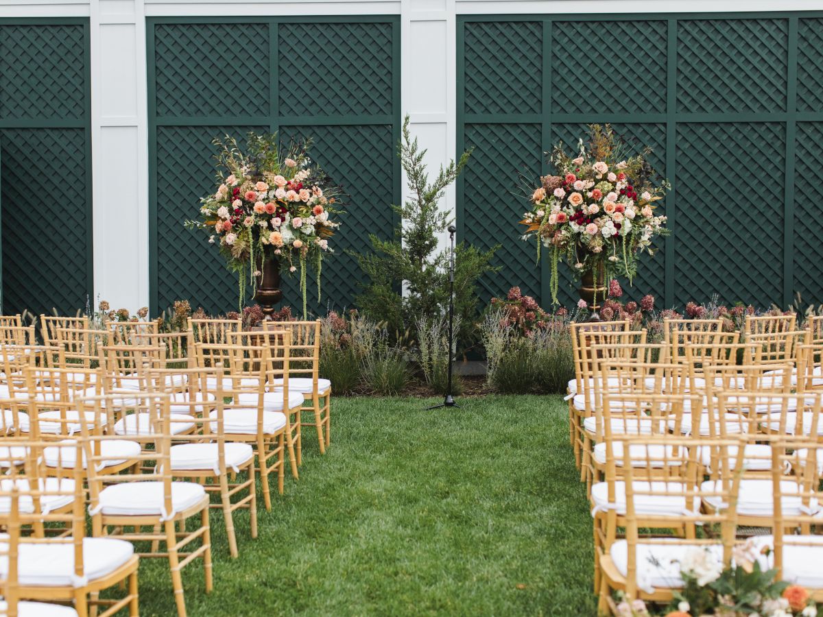 An outdoor wedding ceremony setup with rows of chairs and two large flower arrangements at the front, and a lattice backdrop.