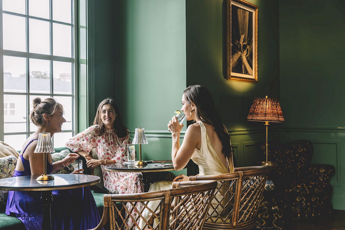 Three women sitting in a stylish, green-painted room with a large window, enjoying drinks and conversation at a small table.