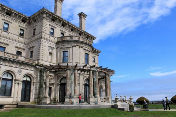 A large, ornate building with classical architecture is seen with a clear blue sky in the background and people walking on the lawn in front of it.