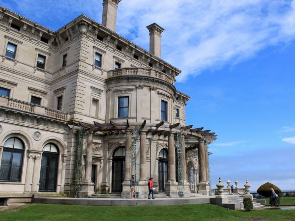 A large, ornate New England mansion with classical architecture, a clear blue sky, and people walking on the front lawn.