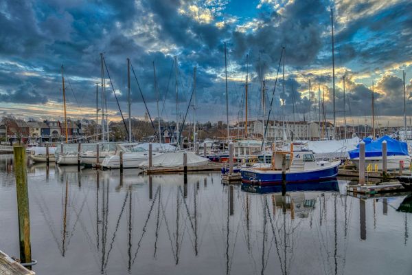 This image shows a marina with sailboats docked, calm water reflecting the boats and masts, and a partly cloudy sky at either dusk or dawn.