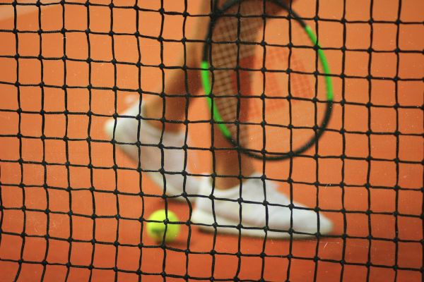 A tennis net, with a racket, ball, and a pair of white shoes visible in the background on a clay court, creating a dynamic sports scene.