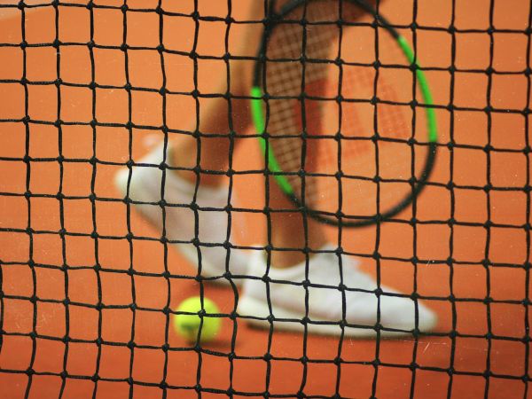 A tennis net, with a racket, ball, and the lower half of a person wearing white tennis shoes standing on an orange tennis court.