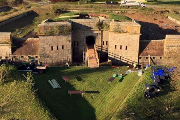 The image shows a well-preserved historic fort with thick stone walls, a central gate, and green lawns, situated in a scenic rural landscape.