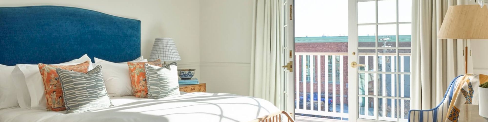 A bright bedroom with a blue headboard bed, wooden bench, striped chair, and French doors leading to a balcony, decorated in a coastal style.