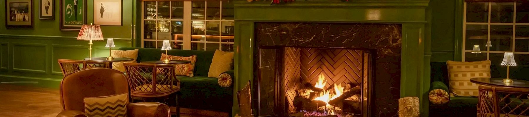 A cozy room with green walls, a lit fireplace, paintings, and comfortable seating, creating a warm and inviting atmosphere with soft lighting.