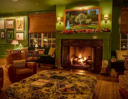 A cozy room with green walls, a lit fireplace, paintings, and comfortable seating, creating a warm and inviting atmosphere with soft lighting.