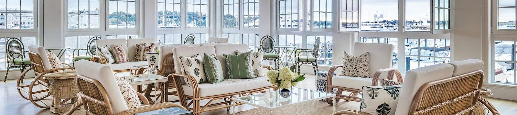 A spacious, bright room with large windows, white furniture with green and neutral cushions, wicker chairs, a rug, and a glass coffee table.