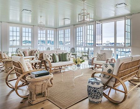 A spacious, bright room with large windows, white furniture with green and neutral cushions, wicker chairs, a rug, and a glass coffee table.