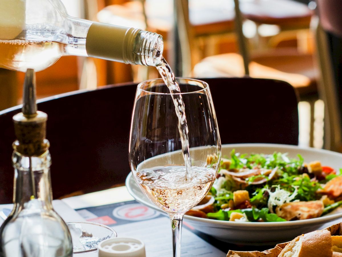 A glass of white wine is being poured at a table set with a salad, bread basket, olive oil, and a pepper grinder in a cozy restaurant.