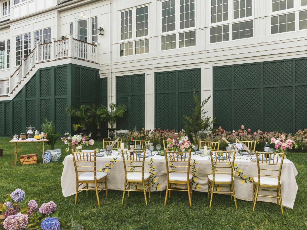 A long dining table set with white tablecloths and gold chairs, floral centerpieces, and an outdoor garden setup in front of a white house with green accents.