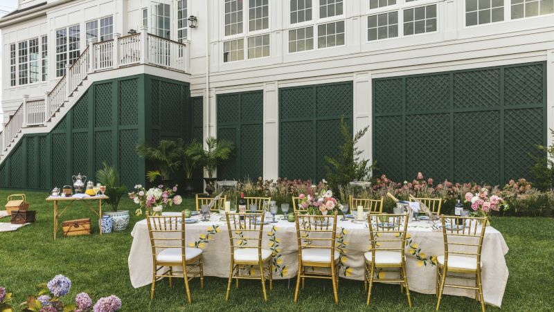 A table set for dining on the lawn in front of a large white building with green trim.