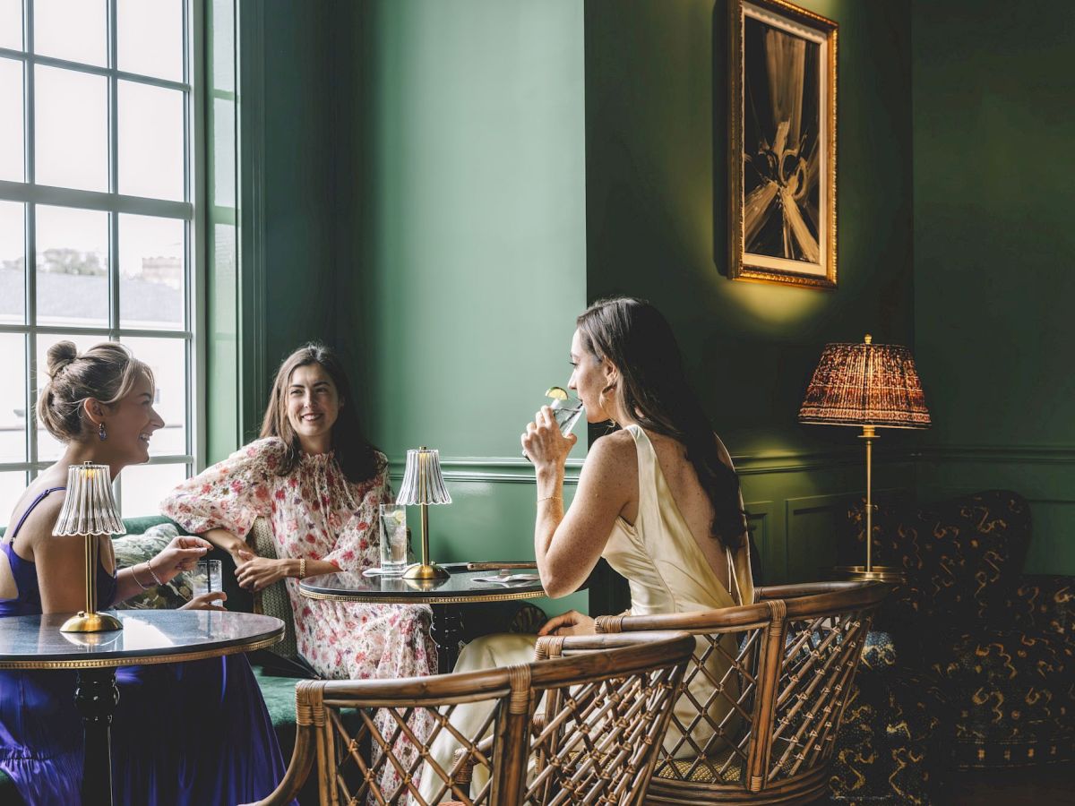 Three women enjoy a bright, cozy lunch at a green-walled cafe, sipping wine and chatting at a glass-topped table by a window.