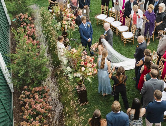 A wedding ceremony taking place in a beautifully landscaped garden with a crowd of people and floral arrangements.