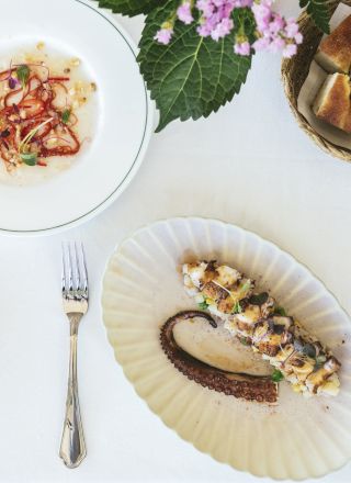 A beautifully set table with a dish of seafood, a garnish, a basket of bread, and a fork and knife, accompanied by decorative plates and a plant.