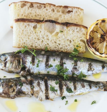 The image shows a plate with two grilled fish garnished with herbs, two slices of bread, and a grilled half of lemon.