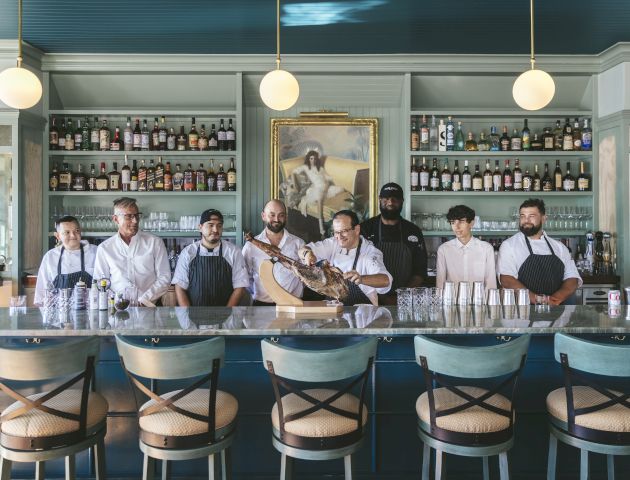 A group of people in chef uniforms stands behind a counter in a bar or restaurant with a painting and shelves of bottles in the background.