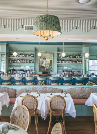 The image shows an elegant restaurant with white tablecloths, cane chairs, chic decor, a wall bar, and a painting in the background.