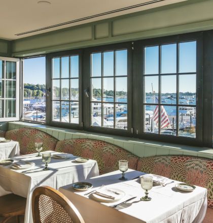 The image shows a cozy restaurant interior with tables set for dining. Large windows offer a scenic view of a marina with boats docked.