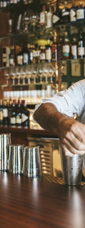 A bartender with white hair and beard is preparing a drink at a bar with various bottles and shakers lined up on the counter.