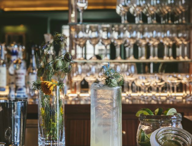 A bar with shelves of liquor bottles and wine glasses, a cocktail garnished with herbs, a vase with flowers, and a lime-filled jar on the counter.