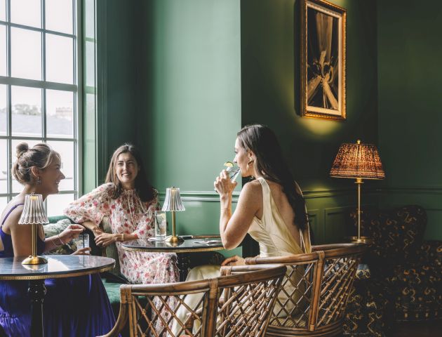 Three women are sitting and chatting in a stylish, green-painted room with elegant furniture and a framed painting on the wall.