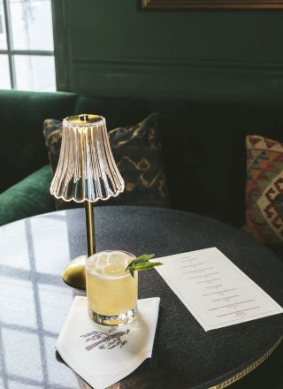 A stylish table setup with a lamp, a cocktail garnished with lime and mint, a napkin, and a menu in a cozy room with green walls and a cushioned seat.