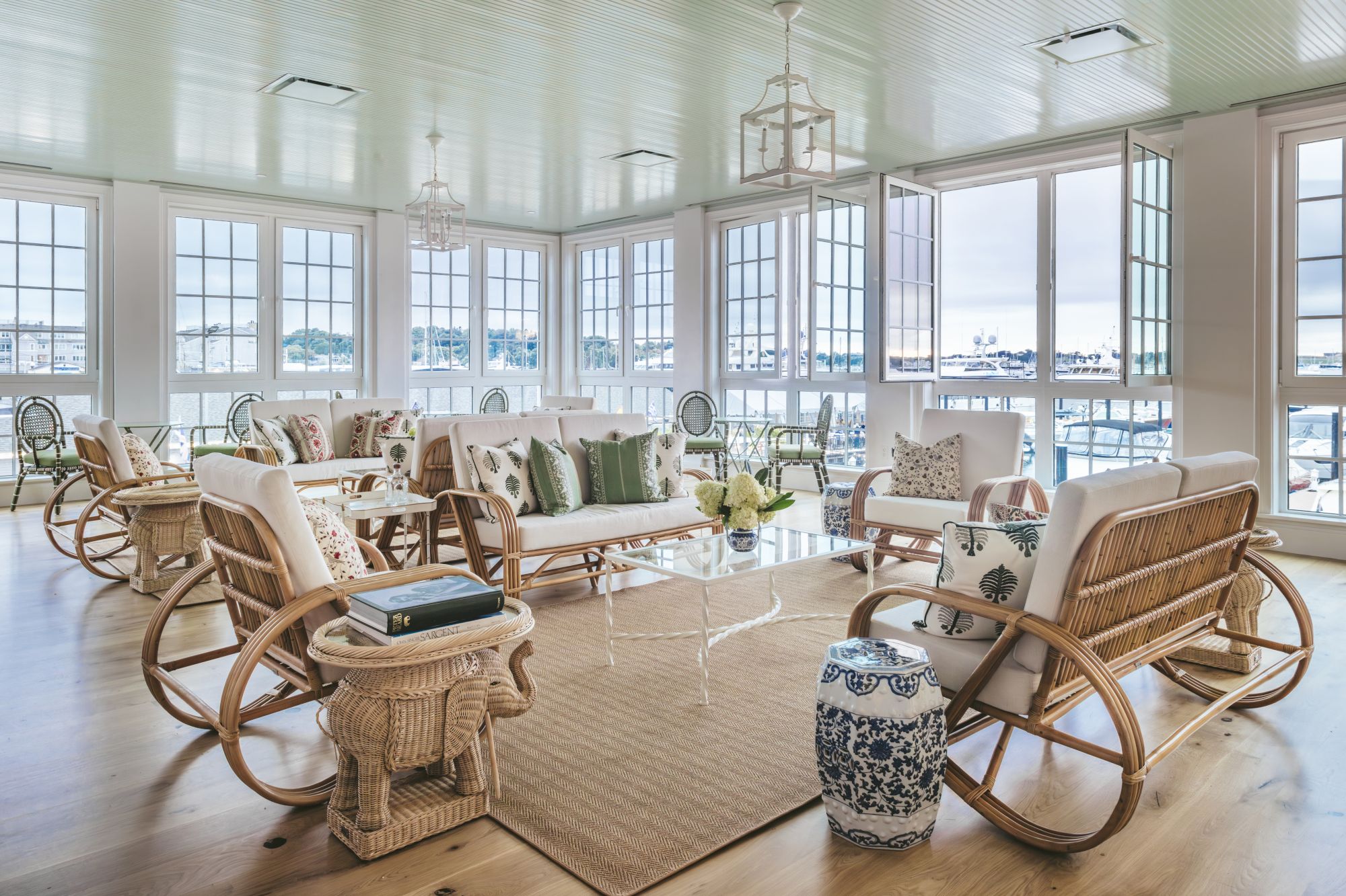 A bright, spacious room with large windows and wicker furniture, featuring a rug and decorative pillows, overlooking a serene waterfront.