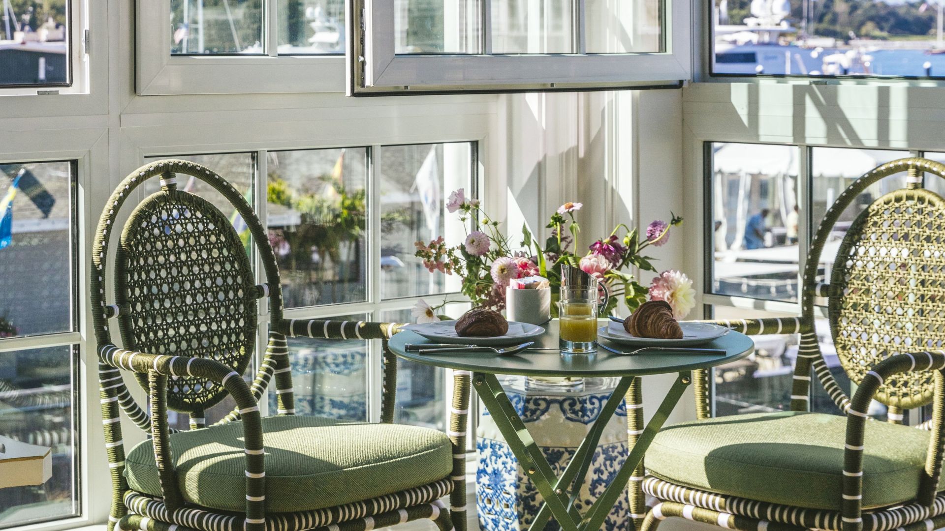 A cozy dining setup for two with wicker chairs, a round table, pastries, juice, and flowers, situated by large windows with a scenic view.