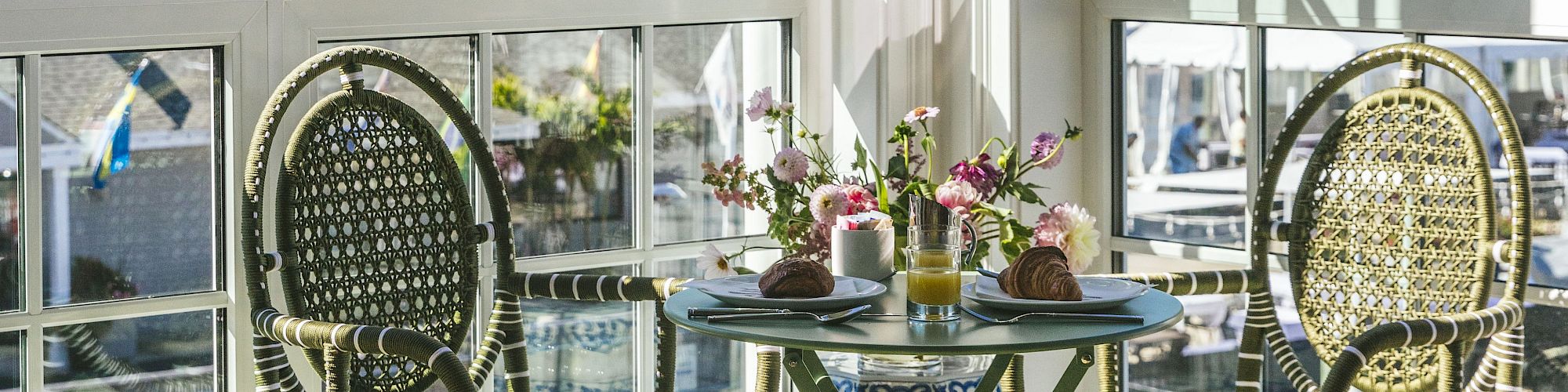 A cozy dining setup for two with wicker chairs, a round table, pastries, juice, and flowers, situated by large windows with a scenic view.