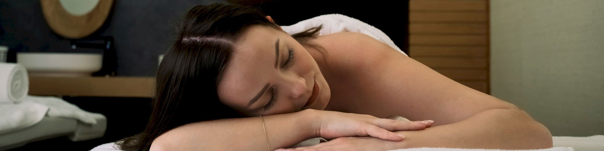 A person is lying down on a massage table, looking relaxed with eyes closed, in what appears to be a spa setting.