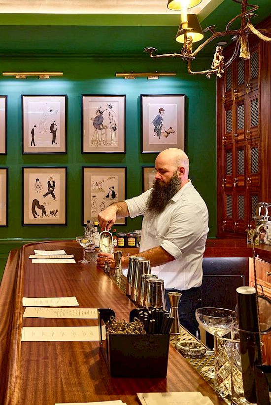 A bearded man is mixing a drink behind a wooden bar in a classy setting with green walls and framed pictures.