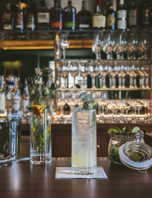 A stylish bar with bottles, glasses, and a cocktail on the counter. Decor includes flowers in a vase and garnishes on the side.