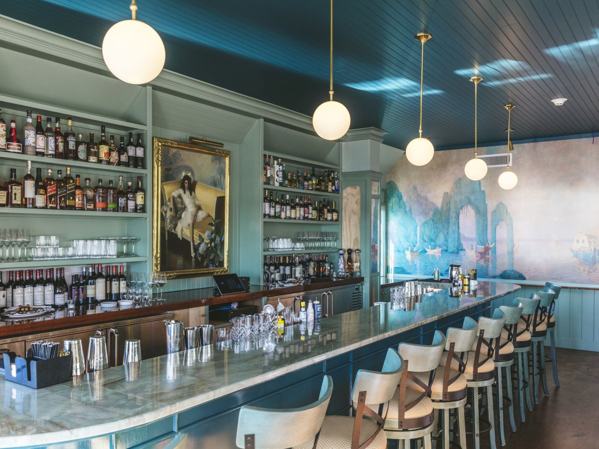 A stylish bar at Flora with a long counter, stools, shelves of bottles, a painting, globe lights, and a mural on the back wall.