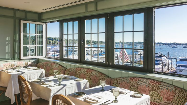 This image shows a cozy restaurant interior with tables set for dining, large windows offering a scenic view of boats on the water.