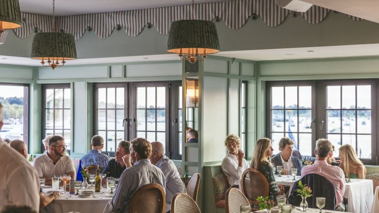 The image shows a busy restaurant with people dining at tables, large windows with views, and elegant decor featuring green accents and hanging lights.