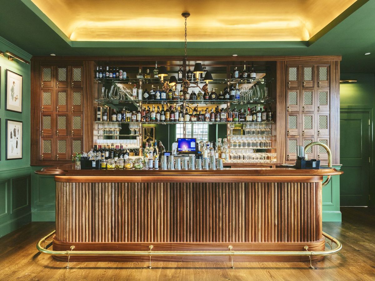 A stylish bar at Studio Bar with a long wooden counter, shelves of bottles, a chandelier, and green walls adorned with paintings.