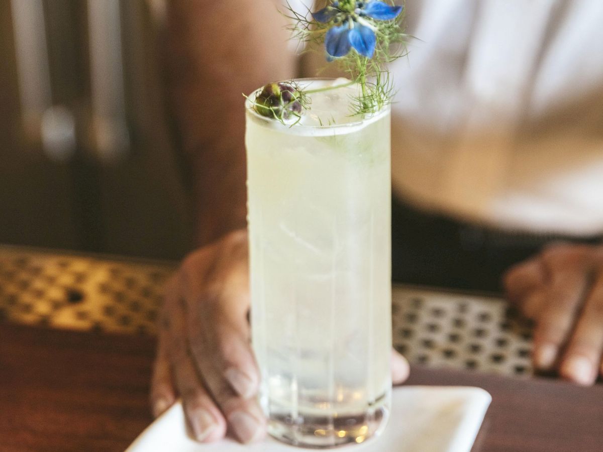 A close-up view of a tall glass of a light-colored cocktail garnished with a blue flower on a napkin, set down by a bartender.