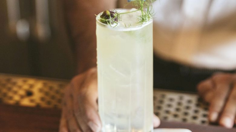 A bartender places a tall glass of a light-colored cocktail garnished with a blue flower on a napkin adorned with a design, on a wooden surface.