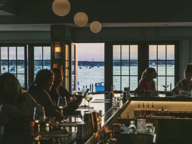 a dimly lit bar with people sitting at the counter and a view of the water with sailboats