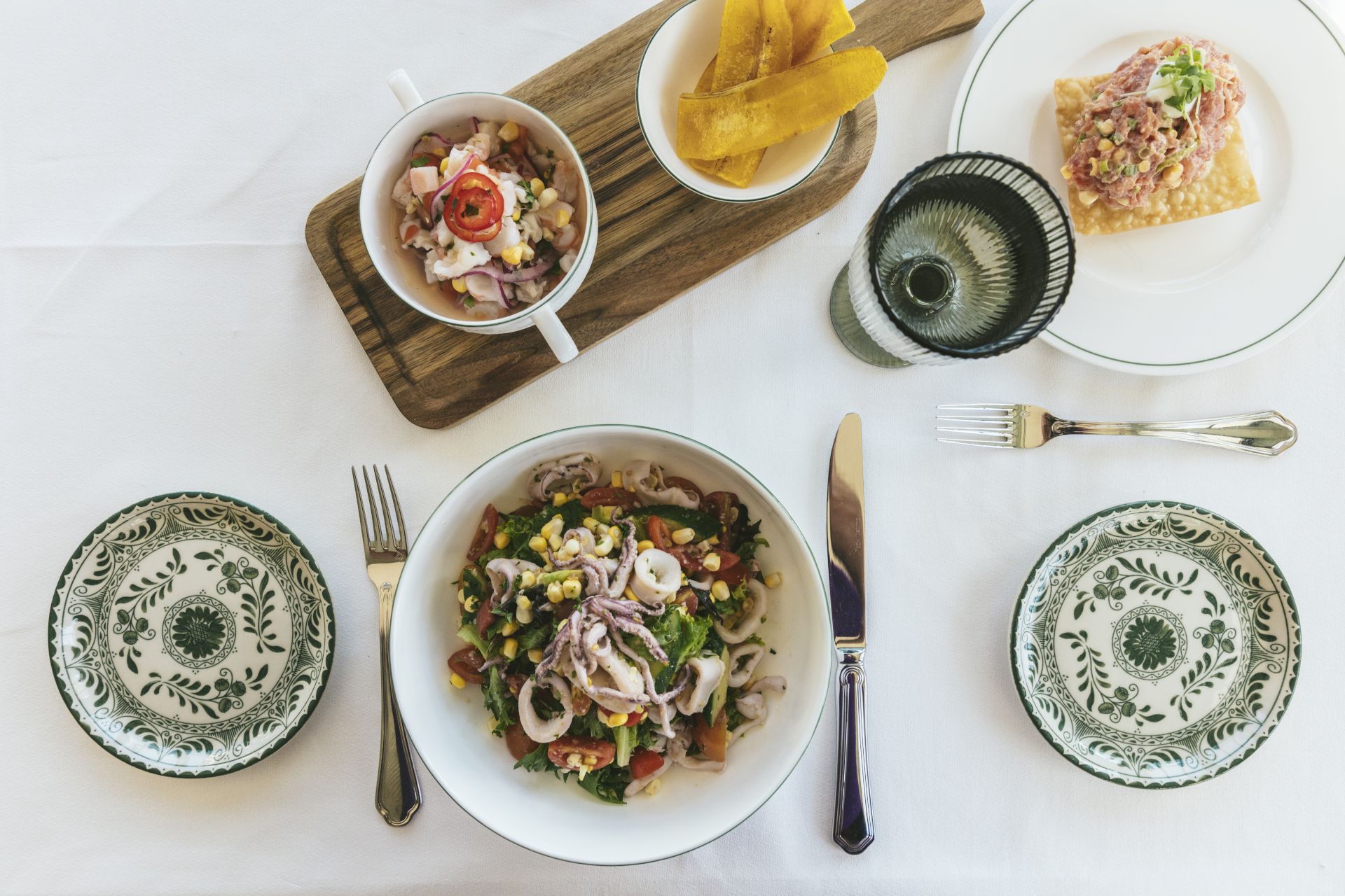 An elegant table setting with two plates of salad, a small bowl of soup, plantain chips, cutlery, and decorative plates and glassware.